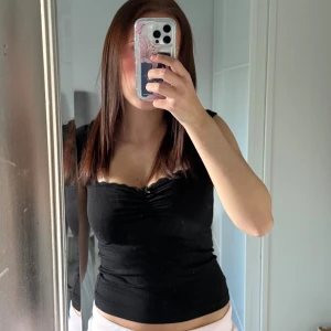 Svart åtsittande linnetopp - Snygg svart linnetopp med smickrande rynkdetalj vid bysten och lätt v-ringad hals. Toppen är ärmlös och har en tight passform som framhäver figuren. Perfekt att matcha med jeans eller kjol för en trendig look.