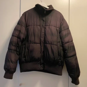 Brun/lila pufferjacka med dragkedja - Säljer en brun/lila pufferjacka med hög krage och dragkedja framtill. Jackan har ribbade muddar vid ärmslut och midja, samt två stora fickor på sidorna. Perfekt för kalla dagar och riktigt skön att bära.❄️💕Den är i 2XL men jag har vanligtvis S/M och jag tycker den sitter lite oversized vilket jag tycker om.