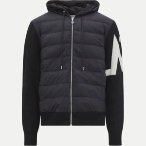 Moncler jacka - Säljer en helt ny svart pufferjacka från Moncler med huva och dragkedja. Jackan har stickade ärmar med en stor vit M-detalj på ena ärmen och Moncler-logga. Kroppen är quiltad och vadderad, perfekt för kyliga dagar. Snygg och sportig look som sticker ut. Jackan är väldigt unik och värmer dig väldigt bra.
