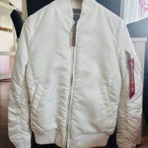 Vit bomberjacka Alpha Industries S - Snygg vit bomberjacka från Alpha Industries med klassisk 'Remove Before Flight'-tag på ärmen. Jackan har dragkedja framtill, ribbade muddar och två fickor med tryckknapp. Tillverkad i polyester med foder och har en cool, clean look som passar perfekt till streetwear.