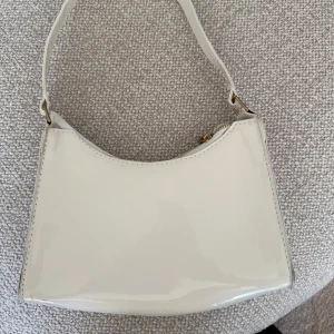 Vit axelväska i glansigt material - Snygg vit axelväska med stilren, minimalistisk design och glansig finish. Väskan har guldfärgade metalldetaljer och dragkedja upptill. Perfekt storlek för det viktigaste och enkel att matcha till olika outfits.