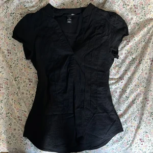 Svart omlottblus - Svart omlottblus från H&M med korta ärmar och lätt v-ringning. Blusen har en figurnära passform och är tillverkad i ett tunt material som ger en elegant känsla. Perfekt att styla med jeans eller kjol för en snygg look.