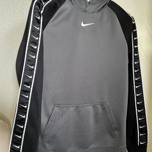 Grå och svart Nike hoodie XL - Snygg grå och svart hoodie från Nike i storlek XL. Tröjan har huva, lång ärm och en stor magficka. Ärmarna har svarta paneler med vita Nike-loggor längs sidorna och en vit piping. Perfekt för chill eller träning.