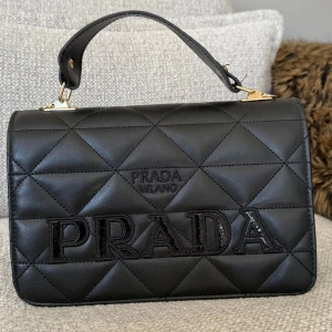 Väska  - Snygg svart handväska med stort broderat logotyp framtill och guldfärgade metalldetaljer. Väskan har både handtag och avtagbar axelrem med logga. Tillverkad i skinn och har en lyxig känsla med klassisk design.