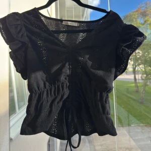 Svart blus med volangärm  - Supersöt svart blus  med v-ringning, volangärmar och snörning framtill. Blusen är croppad och har broderade detaljer i tyget som ger en luftig känsla. Perfekt för dig som gillar feminina och trendiga toppar.