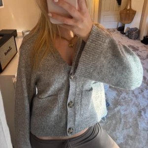 Grå stickad kofta med knappar - Mysig grå stickad kofta med stora knappar framtill och två fickor. Koftan har v-ringning, breda ärmar och ribbade muddar. Perfekt att slänga över en topp för en chill look.