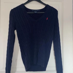 Mörkblå stickad tröja Ralph Lauren - Mörkblå kabelstickad tröja från Ralph Lauren Sport med röd broderad logga på bröstet. V-ringad modell med långa ärmar och ribbade muddar. Perfekt för lager-på-lager och snygg till jeans eller kjol.