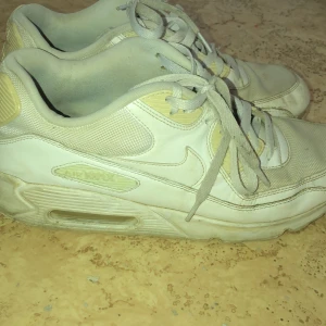 Nike Air Max vita sneakers - Nike Air Max sneakers i vit färg med ljusgula detaljer. Klassisk låg modell med snörning och synlig Air Max-dämpning i sulan. Ovandel i mesh och syntet, med rund tå och platt sula. Perfekt för dig som gillar retrostil och streetwear.