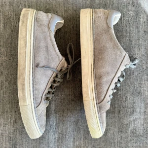 Grå mockasneakers från J.Lindeberg - Snygga grå sneakers från J.Lindeberg med ovandel i mocka och detaljer i ljusgrått skinn vid hälen. Skorna har platt sula, rund tå och klassisk snörning. Perfekta för dig som gillar stilrena och neutrala färger.