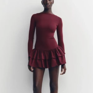 Vinröd volangklänning från Zara - Vinröd kort klänning från Zara med lång ärm och volangkant nedtill. Klänningen har en figurnära passform och är tillverkad i ett mjukt material som sitter bekvämt. Klänningen är stretchig så passar Xs-M. Perfekt nu till hösten!🍁