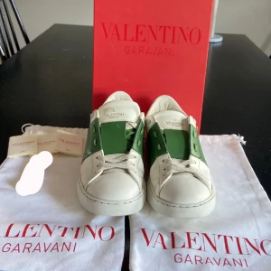 Valentino Garavani Open sneakers vit/grön - Snygga Valentino Garavani Open sneakers i vitt skinn med grön panel och ikoniska nitar på hälen. Klassisk rund tå, platt sula och snörning. Kommer med originalpåse och box. Perfekta för dig som vill sticka ut med lyxig streetstyle. Välj mellan 2 boxar en vit eller röd, allt på bilden följer med även en valentino påse att lägga skorna i! Skorna är sönder vid tårna kan lämna in och fixa men då gäller 2300 eller så köper ni skorna när dem i sönder för 1500 Borde kosta runt 400 och laga 