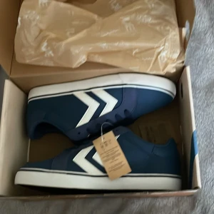 Blå sneakers från Hummel storlek 39 - Snygga mörkblå sneakers från Hummel med klassiska vita pilar på sidorna. Skorna har snörning, vit sula och är tillverkade i syntetmaterial. Perfekta för dig som gillar stilren och sportig design med dansk touch.