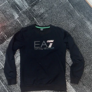 Svart EA7 långärmad tröja - Svart långärmad tröja från EA7 Emporio Armani med tryckt logga i silver och grått på bröstet. Tröjan har rund halsringning med en grön detalj på insidan av kragen. Perfekt för dig som gillar stilrena märkesplagg.