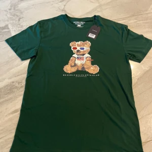 Grön t-shirt med nalle från Polo Club - Snygg grön t-shirt från Beverly Hills Polo Club i 100% bomull. Framsidan har ett coolt tryck med en pixel-art nalle i 3D-glasögon och klubbens logga under. Regular fit med klassisk rund hals och korta ärmar. Perfekt för dig som gillar streetstyle och unika prints.