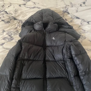 Svart pufferjacka från Calvin Klein Jeans - Säljer en svart pufferjacka från Calvin Klein Jeans med huva och dragkedja framtill. Jackan har diskret CK-logga på bröstet och två fickor med dragkedja. Perfekt för kyliga dagar och riktigt snygg till jeans. Jackan har haft ett hål som nu är fixat💕 köpte för 2499kr 