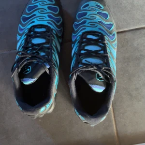 Nike TN Air sneakers blå och svart - Knappt använda Nike Air sneakers i blått och svart med vågigt mönster och svarta snören. Skorna har meshdetaljer för extra andningsförmåga och en sportig vibe. Perfekta för dig som gillar att sticka ut med cool design och bekväm passform.