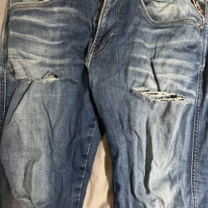 Blå Replay jeans med slitningar - Säljer ett par blå Replay jeans med slitna detaljer över låren och klassisk femficksdesign. Jeansen har snygga kontrastsömmar och broderad logga på bakfickan. Perfekt för dig som gillar en avslappnad och trendig look.