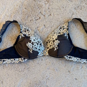 Mörkblå spets-bh från Intimissimi - Säljer en mörkblå bh från Intimissimi med vacker vit spets och blommiga broderier. Bh:n har tunna axelband och genomskinliga mesh-detaljer som ger en lyxig känsla. Perfekt för dig som gillar eleganta underkläder med fina detaljer.