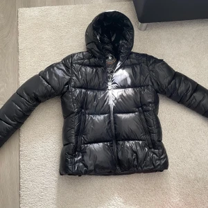 Svart pufferjacka från Save The Duck - Säljer en svart, glansig pufferjacka från Save The Duck med huva och dragkedja. Jackan är som ny.  