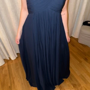 Mörkblå långklänning med v-ringning - Elegant mörkblå långklänning med v-ringad omlott-topp och fint fallande kjol. Klänningen har breda axelband och ett plisserat liv som ger en snygg siluett. Perfekt för dig som vill ha en klassisk och stilren look.