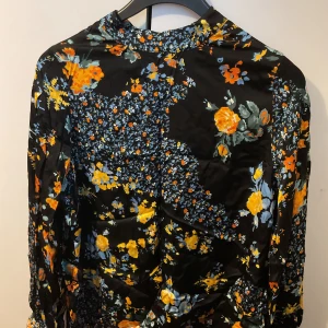 Svart blommig långärmad blus - Svart blus med färgstarkt blommönster i gult, orange och blått. Långärmad modell med hög hals och mjukt, glansigt tyg som ger en lyxig känsla. Perfekt för dig som vill sticka ut med en cool och trendig look.