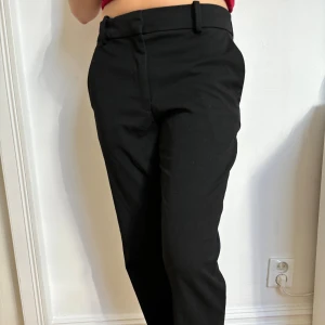 Lågmidjade svarta kostymbyxor med raka ben - Använda två gånger. Svarta kostymbyxor med klassisk rak passform och bälteshällor. Byxorna har diskreta fickor bak och stängs med dragkedja och knapp framtill. 