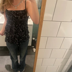 Svart spetslinnetopp från Kappahl - Supersöt svart linne-topp från Kappahl med tunna axelband och volangdetaljer. Toppen har ett grått spetsmönster över hela och flera lager volanger som ger en cool och romantisk vibe. Perfekt att styla med jeans eller kjol för en trendig look.