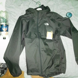 Svart vindjacka från The North Face - Svart vindjacka från The North Face med huva och hel dragkedja framtill. Jackan har en stilren design med logga på bröstet och är tillverkad i ett lätt, vattenavvisande material. Perfekt för blåsiga dagar och enkel att matcha med allt.