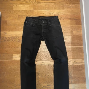 Svarta selvage nudie grim Tim - Snygga svarta jeans från Nudie Jeans Co med klassisk femficksdesign och diskret logga på bakfickan. Jeansen har raka ben och är tillverkade i slitstarkt denimtyg med selvedge-detalj vid benslutet. Perfekta för dig som gillar stilrena och tidlösa jeans. Jeansen är 33:30 i storlek 