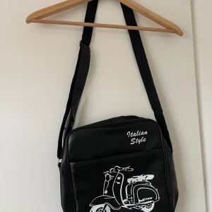 Svart axelväska med scooter-tryck - Cool svart axelväska med vitt tryck av en klassisk italiensk scooter och texten 'Italian Style' på framsidan. Väskan har justerbar axelrem och dragkedja upptill. Perfekt för dig som gillar retrostil och vill sticka ut.