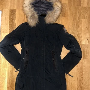 Svart/blå Parajumpers parkas med pälskant - Säljer en svart/blå lång parkas från Parajumpers med fluffig pälskant på huvan och snygga detaljer. Jackan har dragkedja, två stora fickor med dragkedja och broderad logga på ärmen. Perfekt för kalla dagar och riktigt stilren look.