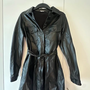 Svart skinnklänning från DeFacto - Cool svart skinnklänning från DeFacto med långa ärmar, knappar framtill och bälte i midjan. Klänningen har bröstfickor med lock och vanliga fickor på sidorna. Perfekt för dig som vill ha en edgy look med snygga detaljer.