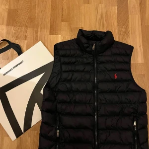 Svart dunväst från Polo Ralph Lauren - Svart dunväst från Polo Ralph Lauren med horisontella quiltade paneler och klassisk ståkrage. Västen har dragkedja framtill, två fickor med dragkedja och den ikoniska röda Polo-loggan broderad på bröstet. Perfekt för lager-på-lager och stilren look.