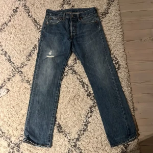 Levi's 501 blå jeans W29 L30 - Klassiska Levi's 501 jeans i blå denim med raka ben och knappgylf. Snyggt slitna med en tydlig reva på vänster lår för en extra edge. Ikonisk läderpatch bak och femficksmodell. Perfekta för dig som gillar vintagevibbar och tidlös stil. Högerfickan är dock trasig men lätt att sy igen!!!
