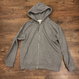 Grå hoodie med dragkedja från Pier One - Grå hoodie från Pier One med hel dragkedja framtill och huva med snörning. Tröjan har långa ärmar, två fickor och är tillverkad i mjuk bomullsblandning. Perfekt för chill dagar eller när du vill ha en enkel och stilren look.