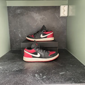 Nike Air Jordan 1 Low svart/röd - Säljer ett par Nike Air Jordan 1 Low med svart och rött läder, vit swoosh och röd yttersula. Klassisk siluett med snörning och rund tå. Perfekta för dig som gillar streetwear och sneakers med ikonisk design.