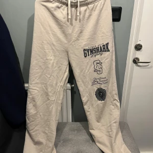 Beiga mjukisbyxor från Gymshark L - Snygga beiga mjukisbyxor från Gymshark i storlek L. Byxorna har tryck med Gymshark-logga och text på ena benet, elastisk midja med snörning och raka ben. Perfekta för chill eller gymmet, tillverkade i mjukt bomullsmaterial.