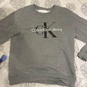 Grå sweatshirt från Calvin Klein Jeans - Säljer en stilren grå sweatshirt från Calvin Klein Jeans med stort svart och vitt logotryck på bröstet. Tröjan har rund hals, långa ärmar och ribbade muddar vid ärmslut och nederkant. Perfekt för en chill och snygg look.