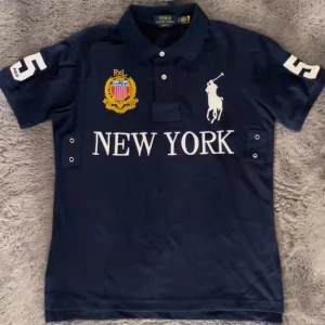 Cheif Keef Polo - Snygg mörkblå t-shirt från Polo Ralph Lauren med vit tryckt text 'NEW YORK' och stort nummer 5 på både ärm och rygg. Klassisk polokrage, broderad logga och emblem i gult och rött framtill. Perfekt för dig som gillar sportig och stilren look.