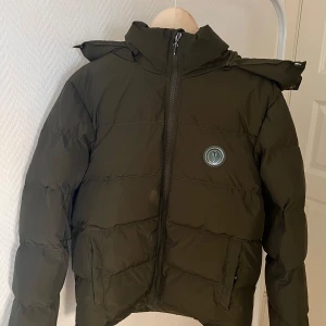 Trapstar grön pufferjacka med huva - Säljer en mörkgrön pufferjacka från Trapstar med stor broderad logga på ryggen och rund patch på bröstet. Jackan har huva med tryckknappar, dragkedja framtill och två fickor. Perfekt för kalla dagar och riktigt snygg streetstil.