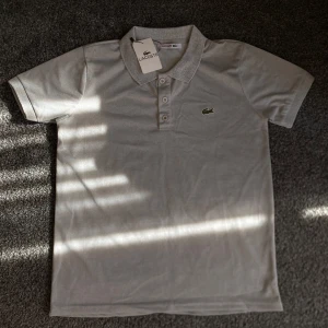 Grå Lacoste piké - Snygg grå t-shirt från Lacoste med klassisk krage och tre knappar framtill. Ikoniska gröna krokodilloggan på bröstet. Tillverkad i mjuk bomull som känns skön mot huden. Perfekt för dig som gillar stilrena och sportiga plagg.