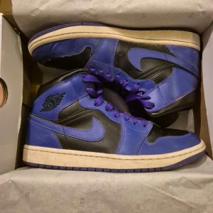 Nike Air Jordan 1 Mid svart/lila - Säljer ett par Nike Air Jordan 1 Mid i svart och lila skinn med klassisk Jordan-logga på sidan. Skorna har lila snörning, vit mellansula och svart/ lila yttersula. Perfekta för dig som vill sticka ut med en ikonisk sneaker med högt skaft och snygg färgkombo. Minimalt creasade med crease skydd på insidan.
