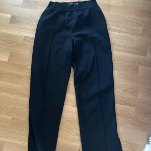 Svarta kostymbyxor från Vero Moda - Svarta kostymbyxor från Vero Moda med elastisk midja och raka ben. Byxorna har en klassisk look med pressveck framtill och är tillverkade i ett mjukt, lätt material som ger en skön passform. Perfekta för en clean och stilren outfit.