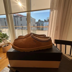 UGG Tasman - Säljer ett par bruna slip-on skor från UGG i mjuk mocka med fluffigt foder och grov sula. Skorna har dekorativt mönster runt öppningen och är perfekta för chill dagar. Skorna är varsamt använda nästintill nyskick utan några defekter. Pris kan diskuteras!