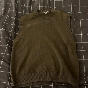 Olivgrön stickad väst med v-ringning - Säljer en olivgrön stickad väst med bred ribbad kant vid hals, ärmhål och nederkant. Västen har en snygg v-ringad design och är loose fit, perfekt att ha över en t-shirt eller skjorta. Enkel och stilren look som funkar till många outfits.