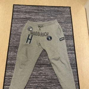 Grå mjukisbyxor från Hoodrich - Snygga grå mjukisbyxor från Hoodrich med flera broderade och tryckta loggor framtill. Byxorna har snörning i midjan och är tillverkade i mjukt bomullsmaterial. Perfekta för en chill och avslappnad stil.