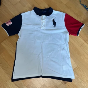 Pikétröja Polo Ralph Lauren - Snygg pikétröja från Polo Ralph Lauren med vit framsida, marinblå rygg och ärm samt röd ärm. Stor broderad logga fram, stor siffra 3 bak och amerikansk flagga på ärmen. Klassisk krage och knappar framtill. Storlek L (14-16) men passar bra för de som vanligtvis bär XS