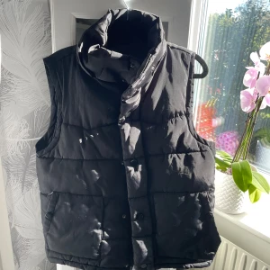 Svart dunväst från Ruosen - Svart quiltad dunväst från Ruosen med hög krage och tryckknappar framtill. Västen har två stora fickor med lock (och en innerficka) och en rak, normal passform. Perfekt att slänga över en hoodie eller långärmad tröja för extra värme.