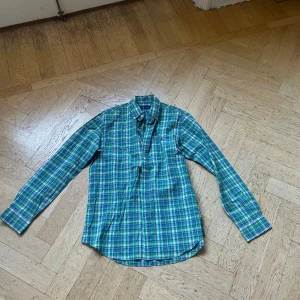 Rutig grön skjorta från Ralph Lauren - Snygg rutig skjorta från Ralph Lauren i grönt, blått och vitt. Klassisk button-down krage, långärmad och med broderad logga på bröstet. Skjortan är i mjuk bomull och har knappar framtill. Perfekt för en chill och stilren look.