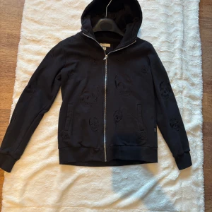 Svart hoodie med dödskallemönster - Svart hoodie från Zara med broderade dödskallar över hela tröjan. Tröjan har dragkedja framtill, huva och långa ärmar med ribbade muddar. Perfekt för dig som gillar edgy streetstyle och vill sticka ut lite extra.